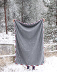 A person holds a gray blanket with flannel trim in front of them in the snow. Snow covered trees and a wooden fence are in the background. 