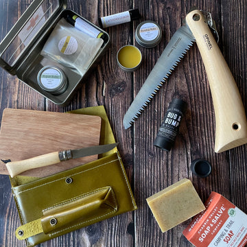 Contents of the Camper Bundle displayed on a wooden surface. A saw, a bar of soap, a knife and board set, a first aid hit, and bug and bump salve.