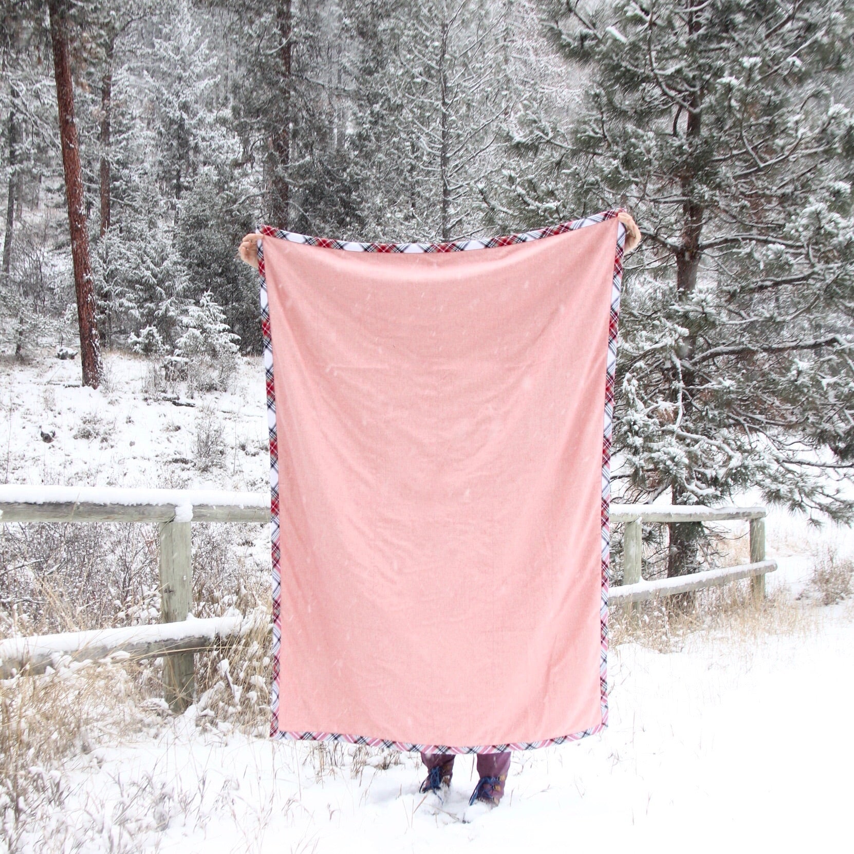A person holds a pink blanket with flannel trim in front of them in the snow. Snow covered trees and a wooden fence are in the background. 