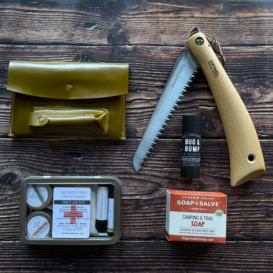 Contents of the Camper Bundle displayed on a wooden surface. A saw, a bar of soap, a knife and board set, a first aid hit, and bug and bump salve.