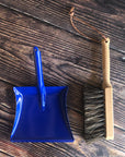 A blue dustpan and wooden brush made for children are placed on a wooden surface. 