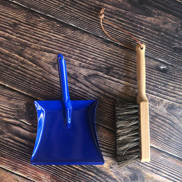 A blue dustpan and wooden brush made for children are placed on a wooden surface. 